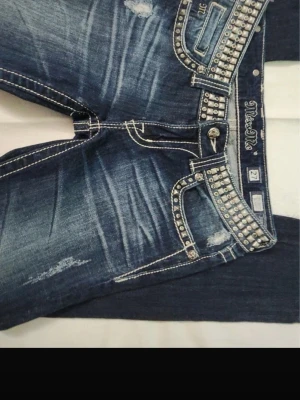 Miss Me blå jeans med nitar - Säljer ett par blå Miss Me jeans med slitningar och coola detaljer som nitar och dekorativa sömmar. Jeansen har låg midja, fem fickor och en snygg tvättad look. Perfekta för dig som gillar statement-plagg och vill sticka ut.