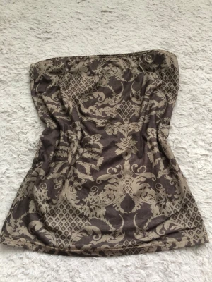 Brun tubtop med barockmönster - Säljer en brun tubtop med beige barockinspirerat mönster. Toppen är axelbandslös och har en mjuk, stretchig passform som sitter snyggt mot kroppen. Materialet känns lätt och bekvämt, perfekt för att styla med jeans eller kjol.
