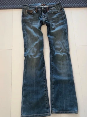 Blå bootcut jeans vintage y2k - Säljer ett par blå bootcut jeans med snygga kontrastsömmar och klassiska fem fickor. Jeansen har låg midja och slitna detaljer vid fickorna för en cool look. Perfekta för dig som gillar retrostil och vill ha ett par jeans med lite vidare ben.