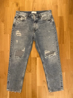 Grå slitna jeans från Calvin Klein w32 - Säljer ett par gråa, raka jeans från Calvin Klein med flera slitningar och hål på framsidan. Jeansen har fem fickor, hög midja och klassisk CK-logga på bakfickan. Tillverkade i denim med vintagekänsla och råa kanter vid benslut.