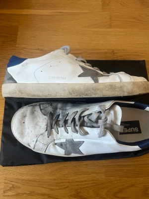Golden Goose vita sneakers med blå detalj - Snygga vita sneakers från Golden Goose med grå stjärna på sidan, grå skosnören och blå detalj på hälen. Skorna har en klassisk låg siluett och är tillverkade i läder med slitna detaljer för en trendig look.