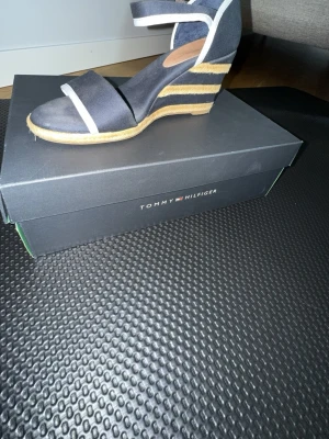 Marinblå kilklackssandal Tommy Hilfiger - Snygg marinblå sandal med kilklack från Tommy Hilfiger. Sandalen har vita detaljer, öppen tå och justerbart ankelband. Klacken är täckt med repdetaljer för en somrig look. Perfekt för dig som vill ha både stil och komfort.