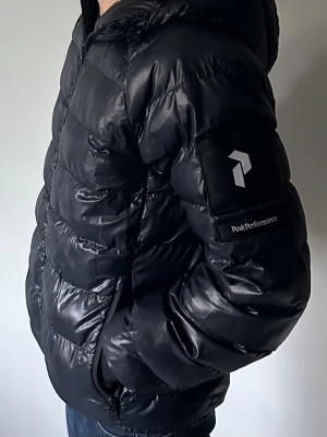 Svart pufferjacka Peak Performance - Snygg svart pufferjacka från Peak Performance med huva och dragkedja framtill. Jackan har en quiltad design, logga på ärmen och praktiska fickor. Materialet är glansigt och ger en modern look. Perfekt för kalla vinterdagar.