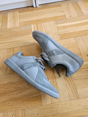 Maison Margiela Gats full Grey - Snygga grå Maison Margiela Gats Paris sneakers med både mocka- och läderdetaljer. Skorna har klassisk låg profil, matchande grå snörning och sula. Perfekta för dig som gillar stilrena och minimalistiska sneakers.