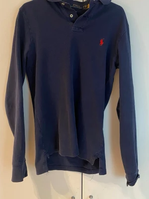 Marinblå långärmad tröja Polo Ralph Lauren - Klassisk marinblå långärmad tröja från Polo Ralph Lauren med krage och två knappar vid halsen. Tröjan har det ikoniska röda Polo-märket broderat på bröstet och är tillverkad i mjukt bomullsmaterial. Perfekt för en avslappnad men ändå stilren look. Finns 3 skavanker som det det finns bild på i slutet lägre pris vid snabb affär