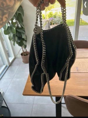 Svart Stella McCartney handväska - Snygg svart handväska från Stella McCartney med silverfärgade kedjedetaljer och rosa tygfoder. Väskan har en mjuk form och kan bäras både i handen och över axeln tack vare de långa kedjehandtagen. Perfekt för dig som gillar trendiga accessoarer.