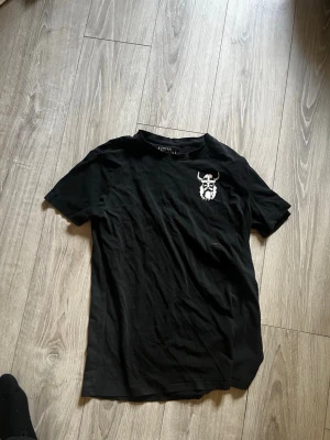 Svart t-shirt från Astungt med tryck - Säljer en svart t-shirt från Astungt i storlek M. T-shirten har ett vitt tryck med vikingamotiv på bröstet och klassisk rund hals. Tillverkad i mjuk bomull och har korta ärmar för en avslappnad look.