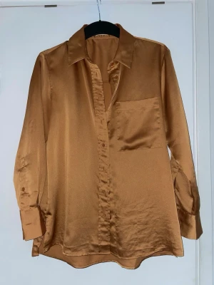 Brun/guld satinskjorta  - Snygg brun/ guldig skjorta i satin med klassisk krage och en bröstficka. Skjortan har långa ärmar med breda manschetter och knappar. 