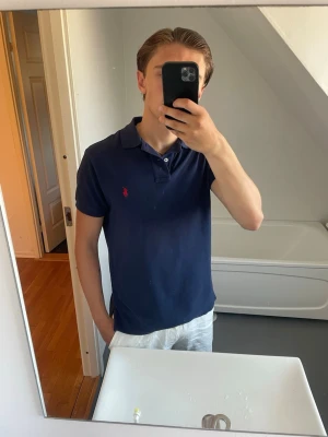 Pikétröja från Polo Ralph Lauren - Klassisk marinblå pikétröja från Polo Ralph Lauren med korta ärmar och krage. Tröjan har den ikoniska röda logotypen broderad på bröstet och är tillverkad i mjuk bomull. Perfekt till jeans eller shorts för en clean och stilren look.
