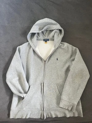 Grå zip hoodie från Polo Ralph Lauren - Snygg grå hoodie med dragkedja från Polo Ralph Lauren. Klassisk design med huva, två fickor framtill och den ikoniska lilla loggan broderad på bröstet. Tillverkad i mjukt sweatshirtmaterial som är perfekt för chill dagar.