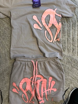 Syna world ser - Säljer ett beige set från Sixth June med matchande t-shirt och shorts. Båda delarna har ett stort rosa grafiskt tryck och shorts med dragsko i midjan. Perfekt för en avslappnad och trendig look.