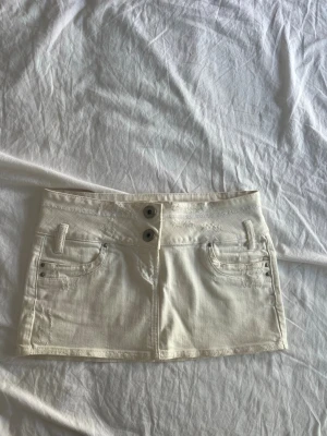 Vit jeanskjol från Self Denim - Snygg vit kort jeanskjol från Self Denim med två knappar framtill och klassiska fickor. Kjolen har en rak passform och är perfekt för dig som gillar en stilren och fräsch look. Passar till många olika outfits och är enkel att matcha.