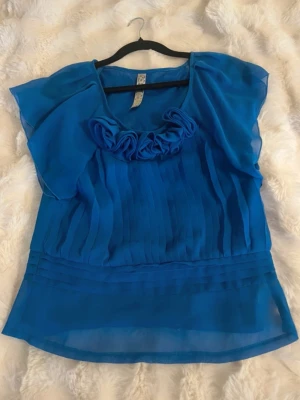 Blå chiffongblus med volangdetalj - Snygg blå blus i luftig chiffong med volangdetalj vid ringningen och tunna, korta ärmar. Blusen har ett lätt transparent tyg och veckade detaljer framtill som ger en cool struktur. 
