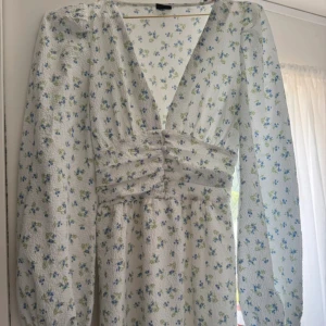Blommig långklänning - Supersöt långklänning från Gina Tricot i vitt med små blå och gröna blommor. Djup v-ringning, långa puffärmar och fina knappar framtill. Klänningen har en lätt strukturerad bomullskänsla och passar perfekt för sommarens alla tillfällen.
