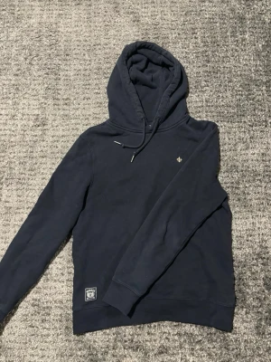 Mörkblå hoodie från Morris - Snygg mörkblå hoodie från Morris med liten logga broderad på bröstet och patch vid nederkanten. Klassisk modell med huva. Tillverkad i mjukt bomullsmaterial som känns skönt mot huden. Perfekt för en avslappnad och stilren look. Bara att höra av sig vid funderingar!