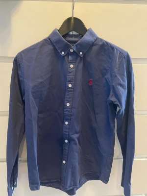 Blå skjorta från Ralph Lauren för barn - Obs! Storlek L för barn. Ca 150-160 cm.            Klassisk blå skjorta från Ralph Lauren med button-down krage och vit knappstängning. Skjortan har lång ärm och den ikoniska röda broderade loggan på bröstet. Tillverkad i bomull och har en tidlös, normal passform.