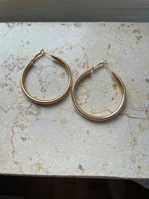 Stora guldfärgade hoops örhängen - Säljer ett par stora, klassiska hoops i guldfärg. Örhängena är runda och har en enkel, stilren design utan dekorationer. Perfekta för dig som gillar tidlösa accessoarer med lite extra attityd. Diameter ca 4-4,5 cm. Aldrig använda.