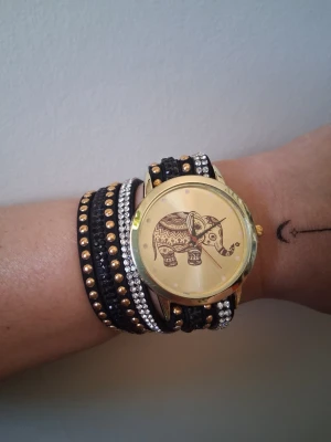Guldig klocka med elefantmotiv - Unik klocka med guldig boett och ett detaljerat elefantmotiv på urtavlan. Armbandet är svart med nitar i guld och silver samt glittrande detaljer som ger en cool och trendig look. Perfekt accessoar för dig som vill sticka ut.
