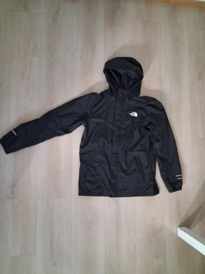 The North Face- Windbreaker - The North Face DryVent Windbreaker Jacket 📏 Kids XL (14/16) – fits like Adult XS / approx. 170cm fit 🌧️ Lightweight waterproof & windproof jacket ✨ Clean matte black colour with embroidered logos front & back Very nice condition overall with only light signs of wear from normal use, nothing major 👌 Name tag inside has been written on previously and covered discreetly. Perfect everyday jacket for streetwear, techwear or outdoor use. Features: • DryVent technology 🌧️ • Adjustable hood • Front zip cl