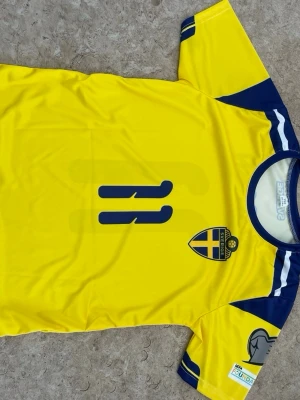 Svensk fotbollströja nummer 11 - Gul fotbollströja från Adidas med blå detaljer, svenska landslagets emblem på bröstet och nummer 11 tryckt både fram och bak. Tröjan har rund halsringning och korta ärmar med blå inslag. Perfekt för dig som vill stötta Sverige på match!