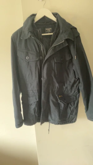 Mörkblå parkasjacka med huva - Snygg mörkblå parkasjacka från Jack & Jones med dragkedja och knappar framtill, flera praktiska fickor och justerbar midja. Jackan har en stor huva och är tillverkad i ett slitstarkt bomullsmaterial. Perfekt för kyliga dagar och enkel att styla till olika outfits.