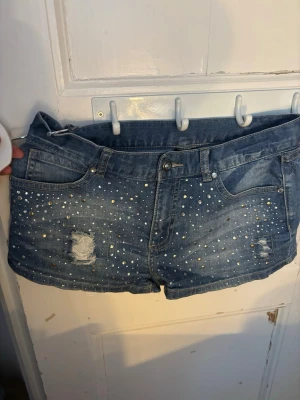 Blå jeansshorts med paljetter - Snygga blå jeansshorts med pärldetaljer framtill och slitna partier för en trendig look. Klassisk femficksmodell med knapp och dragkedja. Perfekta för sommaren och ger en cool vibe till din outfit. Alla hål är synliga i bilden även ett på baksidan