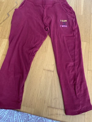 Vinröda mjuka loungewear-byxor - Säljer ett par vinröda loungewear-byxor med fickor och broderad text 'I CAN I WILL' på ena sidan. Byxorna har avslappnad passform och är gjorda i ett mjukt, stretchigt material som passar perfekt för hemmamys eller chill dagar.