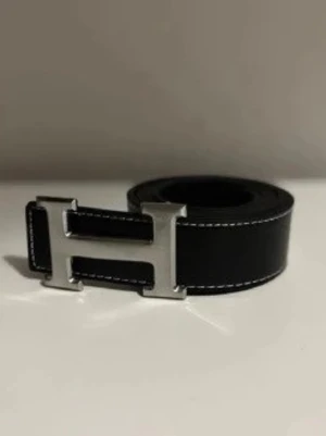 Svart skärp med H-spänne - Snyggt svart skärp med klassiskt H-format metallspänne i silver. Skärpet har vita kontrastsömmar längs kanterna och är gjort i slätt skinn. Perfekt accessoar för att lyfta vilken outfit som helst.