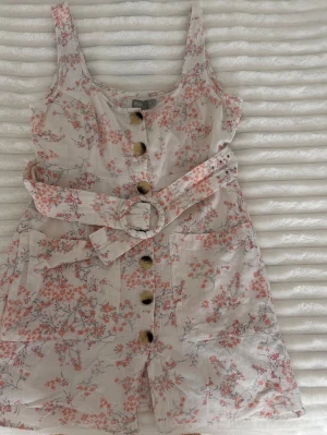 Blommig jumpsuit med bälte - Söt blommig jumpsuit i ljusbeige med rosa och röda blommor. Har breda axelband, stora knappar framtill och ett matchande tygbälte med rund spänne. Två fickor fram och korta ben. Perfekt för sommardagar!