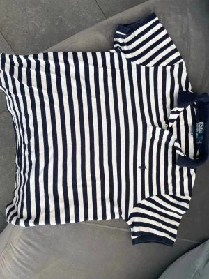Randig pikétröja från Polo Ralph Lauren - Säljer en klassisk kortärmad pikétröja från Polo Ralph Lauren med marinblå och vita ränder. Tröjan har krage, knappar vid halsen och en liten broderad logga på bröstet. Perfekt för en avslappnad men ändå stilren look.