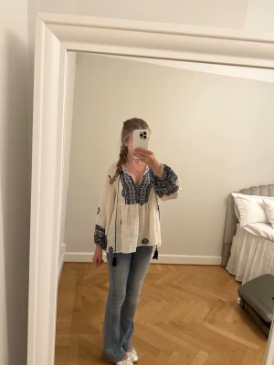 Vit boho-blus med blå brodyr - Säljer en luftig vit blus med blå broderade detaljer och tofsar vid halsen. Blusen har v-ringning, långa ärmar med resår vid handlederna och ett avslappnat, flowy snitt. Perfekt för en avslappnad och trendig look . Använd en gång 