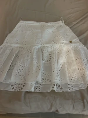 Vit volangkjol med broderade detaljer - Söt vit kortkjol med volang och broderade hålmönster. Kjolen har en luftig känsla och passar perfekt till varma dagar. Tillverkad i bomull för en bekväm och lätt känsla.