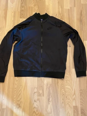 Svart bomberjacka från Nike - Snygg svart bomberjacka från Nike med diskret broderad logga på bröstet. Jackan har dragkedja framtill, ribbade muddar och krage samt lång ärm. Perfekt för dig som gillar sportig och clean stil.