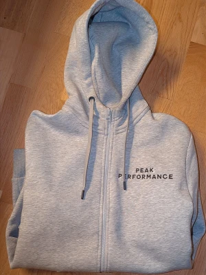 Grå hoodie Peak Performance - Snygg grå hoodie från Peak Performance med dragkedja framtill och svart logga på bröstet. Tröjan har huva med dragsko och två stora fickor framtill. Tillverkad i mjukt sweatshirtmaterial, perfekt för en avslappnad stil. Helt ny testad hemma 2-3 ggr helt ny köpt stadium för ett tusen kr 