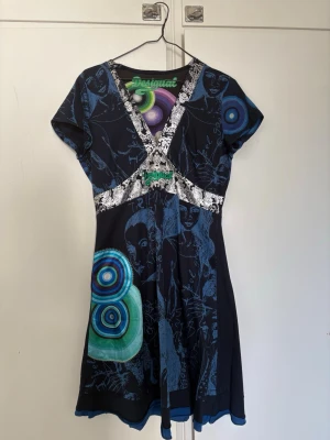Mönstrad midi från Desigual - Unik midiklänning från Desigual i mörkblått med färgstarka cirklar och grafiskt mönster. V-ringad med kontrasterande vita detaljer och korta ärmar. Materialet känns mjukt , troligen bomull och polyesterblandning. Perfekt för dig som gillar statement-plagg.
