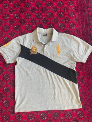 Grå pikétröja från Polo Ralph Lauren - Snygg grå pikétröja från Polo Ralph Lauren med svart diagonal rand och broderade detaljer i gult och rött på bröstet. Klassisk krage och korta ärmar. Perfekt för en sportig och stilren look.