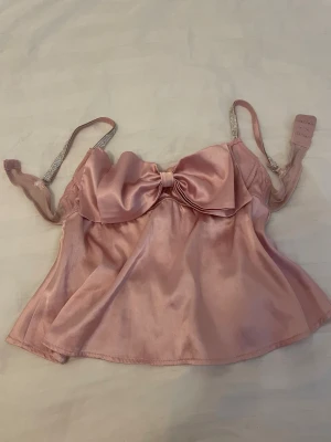 Rosa satin body med rosett och strassband - Supersöt rosa topp i glansigt satinmaterial med stor rosett framtill och smala axelband dekorerade med strass. Urringad modell med lätt volang nertill och hakknäppning bak. Perfekt för dig som vill ha något extra fint i underklädeslådan eller ha som en fin fest topp.