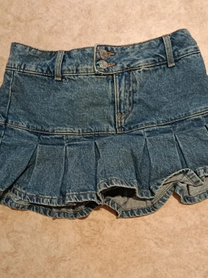 Jeansskort med volangkant - Snygg jeansskort i klassisk blå denim med volangkant nertill och dubbla knappar framtill. Skorten har bälteshällor och fickor fram, samt shorts under kjoldelen. Perfekt för en trendig och avslappnad look.