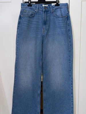 Blå raka jeans från H&M - Säljer ett par klassiska blå jeans från H&M med rak passform och hög midja. Jeansen har fem fickor, bälteshällor och stängs med knapp och dragkedja. Tillverkade i 100% bomull för en skön och avslappnad look. Som du ser på bilden är den helt ny och oanvänd 