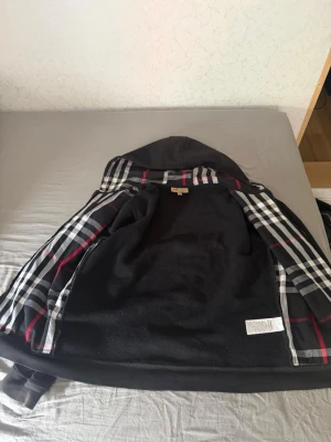 Svart hoodie med dragkedja och rutigt foder - Säljer en svart hoodie från Burberry med dragkedja framtill och klassiskt rutigt foder i huvan och insidan. Hoodien har två fickor fram och ribbade muddar vid ärmslut och nederkant. Perfekt för dig som gillar stilrena detaljer och exklusiv känsla.