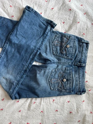 Blå jeans med broderade bakfickor - Säljer ett par blå jeans med snygga broderade detaljer på bakfickorna och silverfärgade knappar. Klassisk femficksmodell i stretchigt denimtyg. Perfekta till en avslappnad look och passar till det mesta i garderoben.