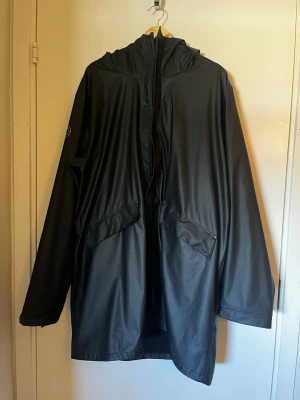 Svart regnjacka med huva - Snygg svart regnjacka med huva och dragkedja framtill. Jackan har två stora fickor med lock och en minimalistisk logga på ärmen. Tillverkad i vattenavvisande material med matt finish och avslappnad passform. Perfekt för regniga dagar.