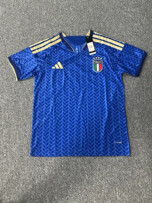 Blå Italien fotbollströja Adidas - Snygg blå Italien fotbollströja från Adidas med gulddetaljer på axlarna och ärmsluten. Tröjan har Italiens landslagsemblem på bröstet, korta ärmar och ett diskret mönster över hela tyget. Tillverkad i lätt och ventilerande material, perfekt för träning eller match.