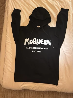 Svart Alexander McQueen hoodie - Snygg svart hoodie från Alexander McQueen med stor vit logga och texten 'EST. 1992' på bröstet. Klassisk känguruficka framtill och justerbar huva med snören. Perfekt för dig som vill ha en streetwear vibe och gillar exklusiva märken.