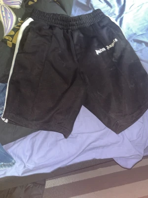 Palm Angels svarta shorts med vita ränder - Säljer ett par svarta shorts från Palm Angels med vita ränder längs sidorna och elastisk midja. Shortsen har loggan broderad framtill och är tillverkade i ett glansigt syntetmaterial som ger en sportig vibe.