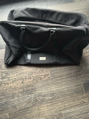 Svart resväska från Hugo Boss - Snygg svart resväska från Hugo Boss med stilren metallbricka framtill. Väskan har två robusta handtag i konstläder och är gjord i slitstarkt tygmaterial. Perfekt för weekendresor eller gymmet, med rymligt huvudfack och dragkedja.