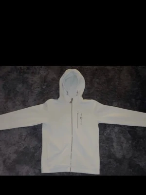 Sail Racing hoodie - Snygg vit hoodie med dragkedja framtill och huva. Hoodien har en bröstficka med dragkedja och långa ärmar. Perfekt för en clean och enkel stil. Materialet ser ut att vara mjukt och bekvämt, troligen bomull eller bomullsblandning.
