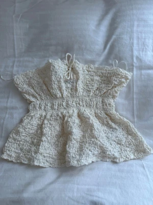 Vit spetsblus från Gina Tricot - Supersöt vit blus från Gina Tricot med volangdetaljer och knyt framtill. Blusen har en smockad midja och är tillverkad i ett spetsliknande material som ger en romantisk vibe. Perfekt till sommaren och lätt att styla med jeans eller kjol.