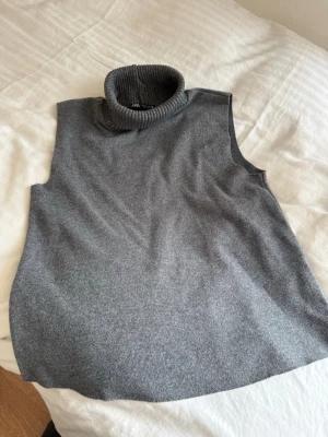 Grå ärmlös polotröja från Zara - Säljer en stilren grå ärmlös polotröja från Zara. Tröjan har en hög, ribbad polokrage och är stickad i mjukt material. Perfekt att bära över en skjorta eller långärmad topp för en trendig lager-på-lager-look.