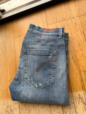 Blå slitna jeans med detaljer - Säljer ett par blå jeans med slitningar och trasiga detaljer framtill. Jeansen har klassisk femficksmodell och en avslappnad passform. Materialet är denim och de har en cool, använd look som passar perfekt till en streetwear-stil. Passar kortare i längd och sitter som 32 i midjan